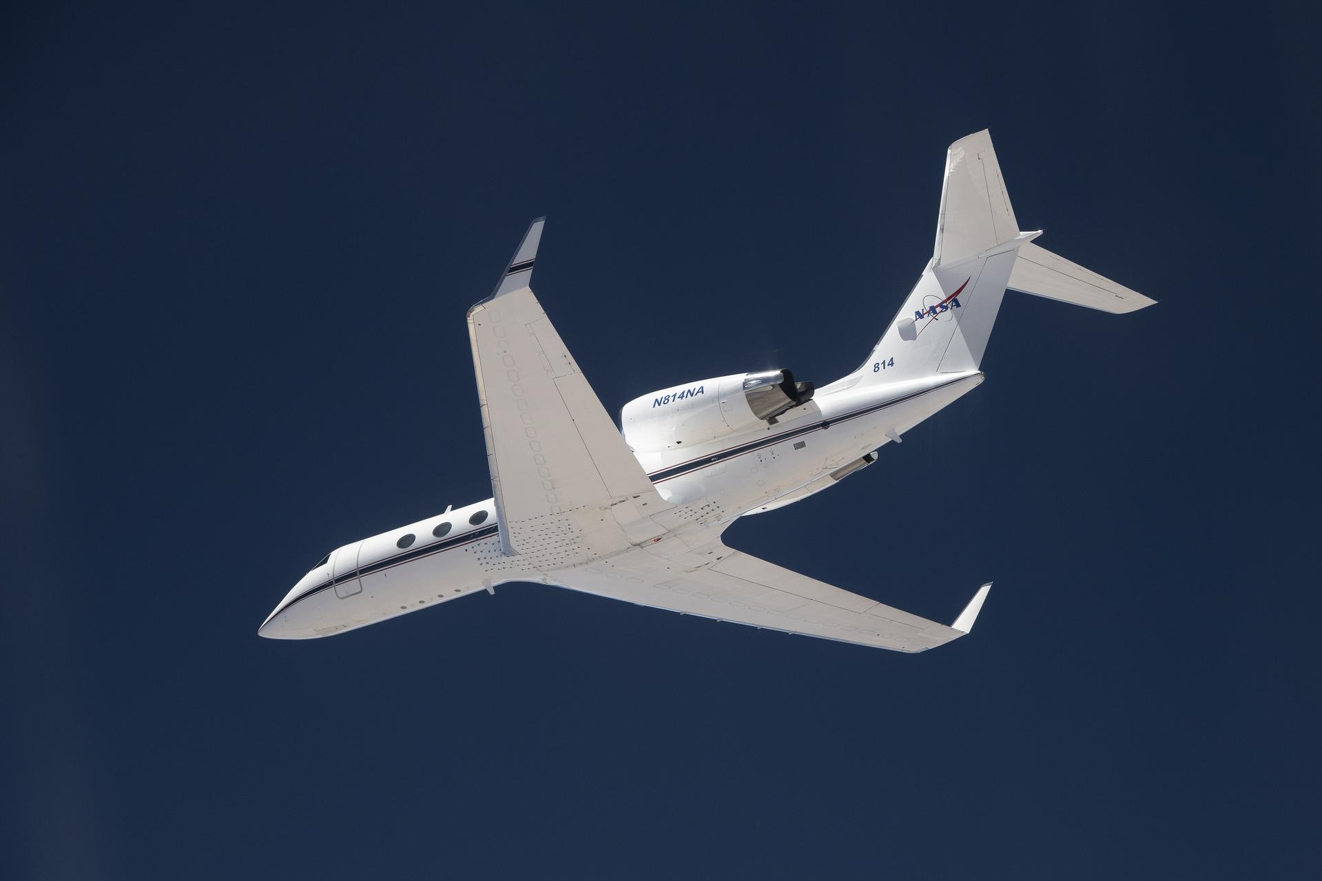 afrc2024 0101 43large.jpg A white plane flies overhead against a dark blue sky. The plane is heading toward the left of the frame, and the NASA insignia is visible on its tail.