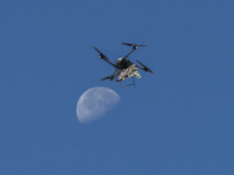 NASA Launches Atmospheric Research Probe A white, blue, and red probe attached to a rotor with four blades flies in the blue sky, just above the Moon.