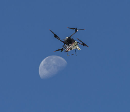 NASA Launches Atmospheric Research Probe A white, blue, and red probe attached to a rotor with four blades flies in the blue sky, just above the Moon.