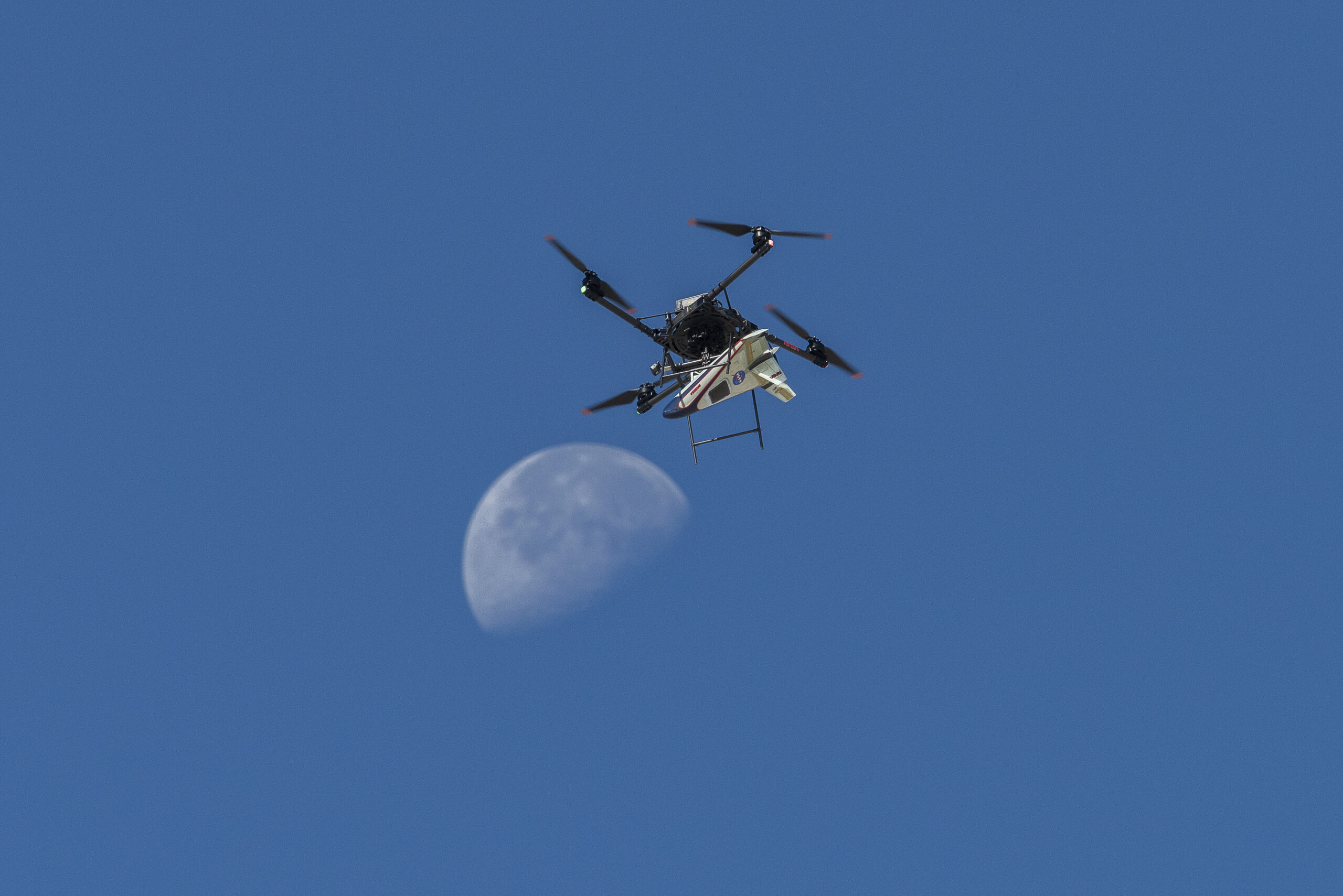 afrc2024 0166 033orig.jpg A white, blue, and red probe attached to a rotor with four blades flies in the blue sky, just above the Moon.