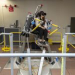 NASA Advances Research on Air Taxi Passenger Comfort A woman sits in a seat with straps atop a metal platform that is tilted to the right side with three base pieces. Her feet rest on a metal platform. Yellow metal bars surround the platform. The woman is wearing black pants and a black and white vest, as well as black virtual reality goggles and a headset.