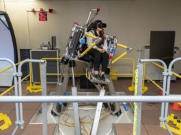 NASA Advances Research on Air Taxi Passenger Comfort A woman sits in a seat with straps atop a metal platform that is tilted to the right side with three base pieces. Her feet rest on a metal platform. Yellow metal bars surround the platform. The woman is wearing black pants and a black and white vest, as well as black virtual reality goggles and a headset.