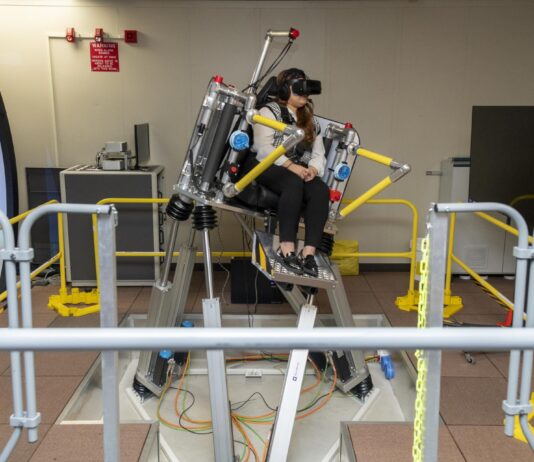 NASA Advances Research on Air Taxi Passenger Comfort A woman sits in a seat with straps atop a metal platform that is tilted to the right side with three base pieces. Her feet rest on a metal platform. Yellow metal bars surround the platform. The woman is wearing black pants and a black and white vest, as well as black virtual reality goggles and a headset.