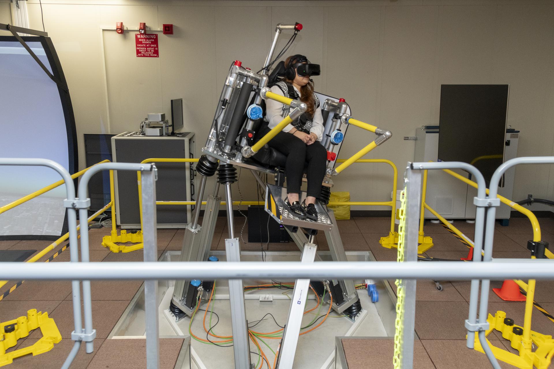 afrc2024 0168 23large.jpg A woman sits in a seat with straps atop a metal platform that is tilted to the right side with three base pieces. Her feet rest on a metal platform. Yellow metal bars surround the platform. The woman is wearing black pants and a black and white vest, as well as black virtual reality goggles and a headset.