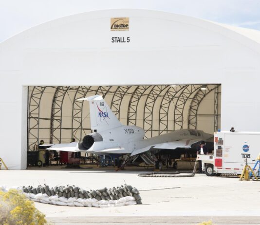 X-59 Engine Ignites, Prepares for Inaugural Flight View from outside a large run stall. The 100-foot-long X-59 sits in the run stall with the engine and the rest of the back of the aircraft sitting outside the run stall’s open bay door.