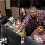 NASA Experts Inspire Students with Perseverance Stories A group of people are sitting at a desk looking at computers with headsets on. A man is pointing and smiling at one of the computer screens.