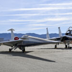 NASA’s X-59 Successfully Finishes Electromagnetic Testing A NASA F-15 aircraft sits 20 feet off the left side of the X-59 aircraft, with a white hangar and hills in the background, during electromagnetic interference testing.