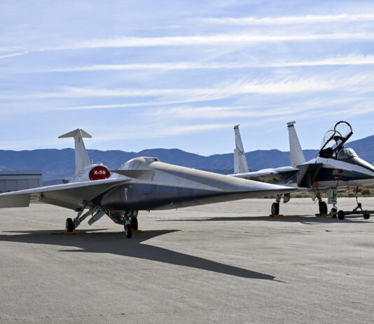 NASA’s X-59 Successfully Finishes Electromagnetic Testing A NASA F-15 aircraft sits 20 feet off the left side of the X-59 aircraft, with a white hangar and hills in the background, during electromagnetic interference testing.