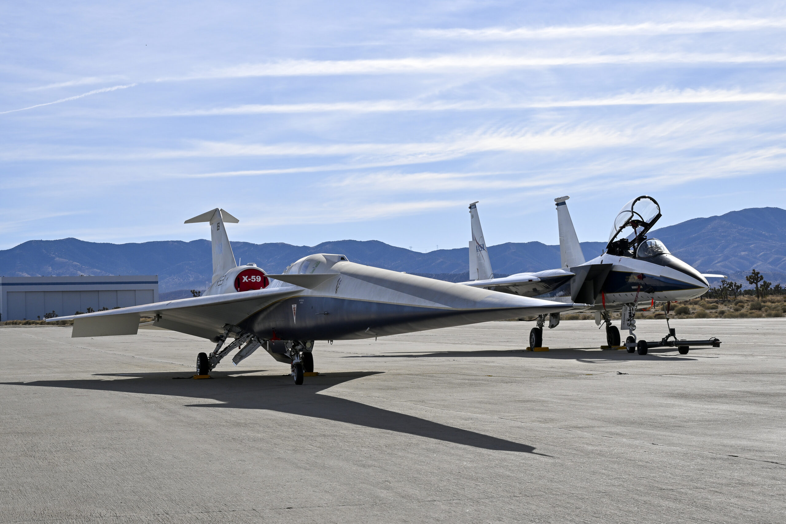 afrc2025 0030 83.jpg A NASA F-15 aircraft sits 20 feet off the left side of the X-59 aircraft, with a white hangar and hills in the background, during electromagnetic interference testing.