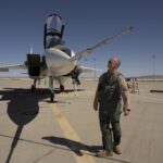 NASA readies second impact sensor for X-59 testing NASA test pilot Nils Larson, wearing a green flight suit and sunglasses, stands at the front of the agency’s F-15D research aircraft, looking up at a long, newly installed shock-sensing probe mounted on the nose. The aircraft is parked on the flightline at NASA’s Armstrong Flight Research Center in Edwards, California, with its canopy open and ladder down.