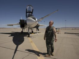 NASA readies second impact sensor for X-59 testing NASA test pilot Nils Larson, wearing a green flight suit and sunglasses, stands at the front of the agency’s F-15D research aircraft, looking up at a long, newly installed shock-sensing probe mounted on the nose. The aircraft is parked on the flightline at NASA’s Armstrong Flight Research Center in Edwards, California, with its canopy open and ladder down.