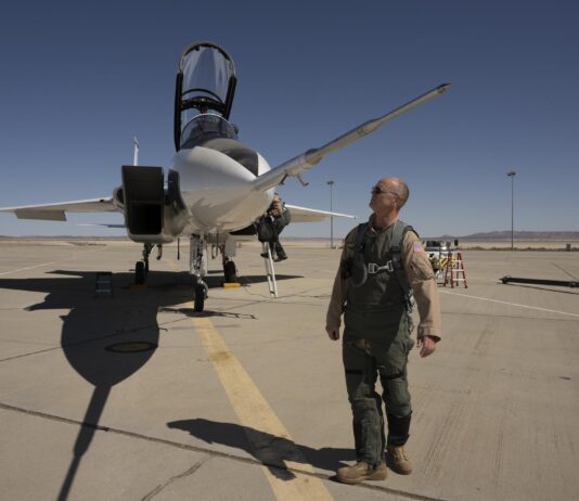 NASA readies second impact sensor for X-59 testing NASA test pilot Nils Larson, wearing a green flight suit and sunglasses, stands at the front of the agency’s F-15D research aircraft, looking up at a long, newly installed shock-sensing probe mounted on the nose. The aircraft is parked on the flightline at NASA’s Armstrong Flight Research Center in Edwards, California, with its canopy open and ladder down.