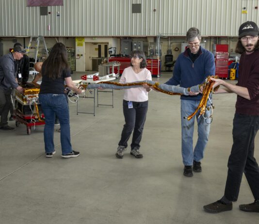 NASA Analyzes Lunar Light to Enhance Earth Monitoring A team of people carry a science instrument across a hangar floor. Two men at the back left of the frame roll a cart carrying the instrument body while four others ahead of the cart walk toward the right of the frame carrying a heavy twine of cables attached to the instrument.
