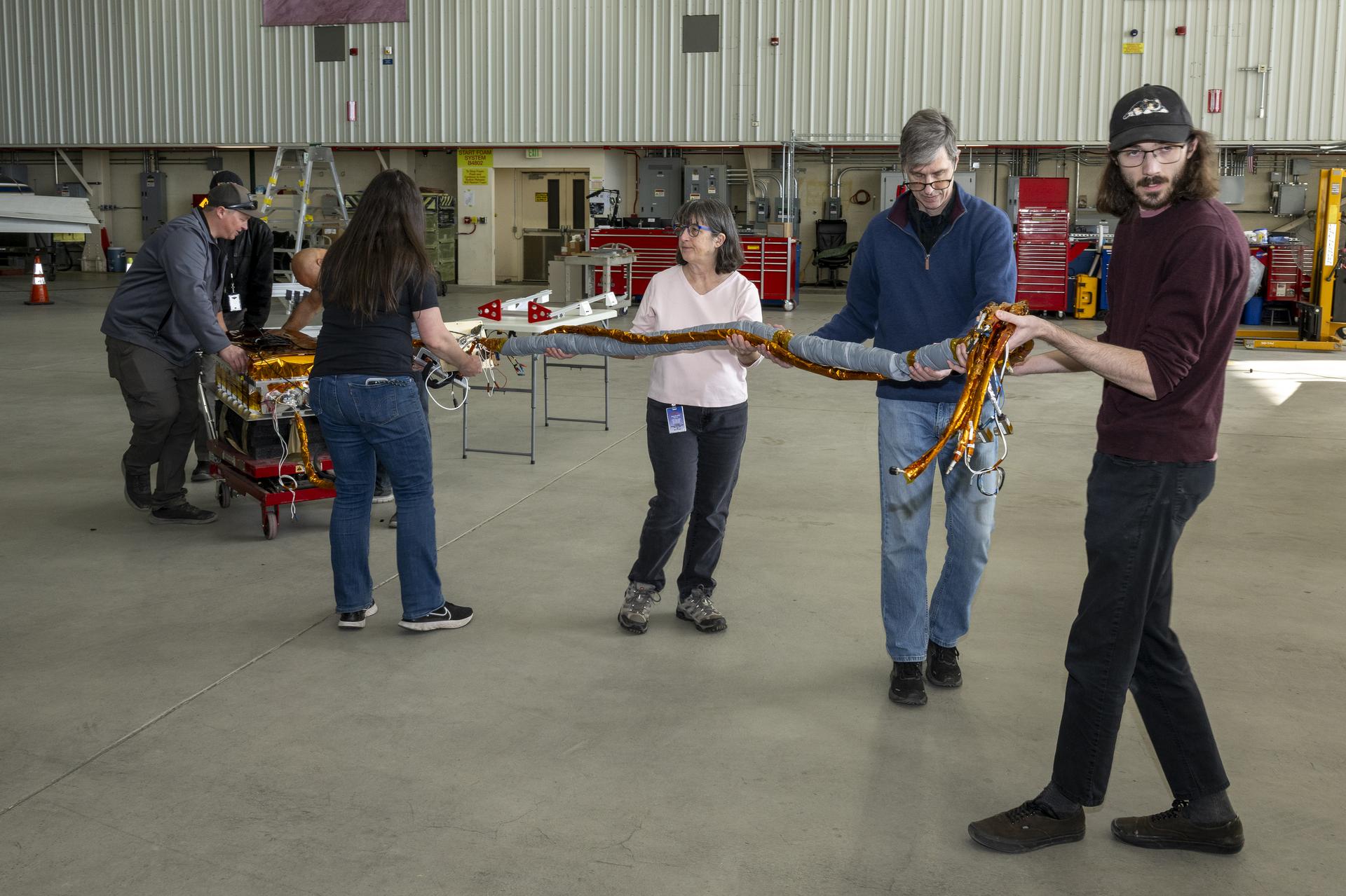afrc2025 0048 063large.jpg A team of people carry a science instrument across a hangar floor. Two men at the back left of the frame roll a cart carrying the instrument body while four others ahead of the cart walk toward the right of the frame carrying a heavy twine of cables attached to the instrument.