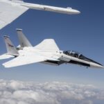 NASA F-15s Test Instruments for Quesst Initiative A NASA F-15D aircraft flies above a cloud layer under a bright blue sky, pitched slightly to the right with its lower right wing closest to the viewer. Two pilots are visible – one in the front seat and one in the rear. The NASA logo appears on the aircraft’s right vertical stabilizer. The aircraft is framed by the wing of another white aircraft in the foreground.