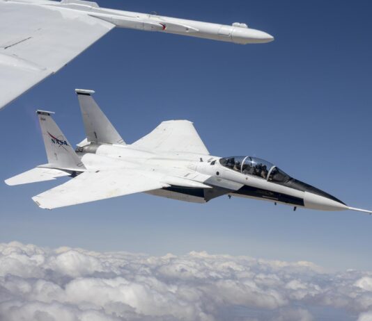 NASA F-15s Test Instruments for Quesst Initiative A NASA F-15D aircraft flies above a cloud layer under a bright blue sky, pitched slightly to the right with its lower right wing closest to the viewer. Two pilots are visible – one in the front seat and one in the rear. The NASA logo appears on the aircraft’s right vertical stabilizer. The aircraft is framed by the wing of another white aircraft in the foreground.