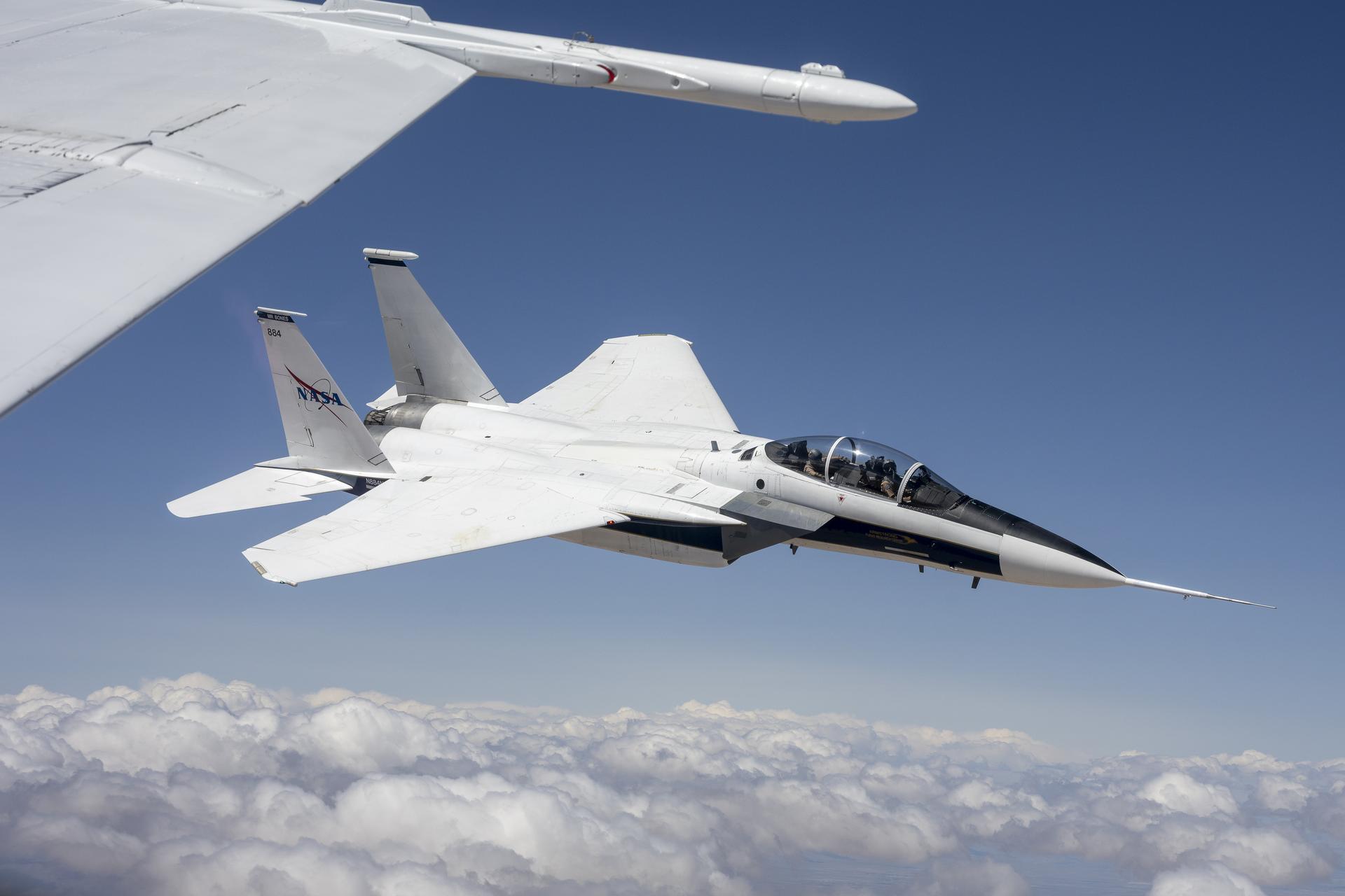 A NASA F-15D aircraft flies above a cloud layer under a bright blue sky, pitched slightly to the right with its lower right wing closest to the viewer. Two pilots are visible – one in the front seat and one in the rear. The NASA logo appears on the aircraft’s right vertical stabilizer. The aircraft is framed by the wing of another white aircraft in the foreground.
