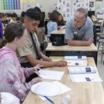 NASA STEM Initiatives Inspire Learning Outside School A group of students sitting at a desk in a classroom work on constructing paper airplanes. A man leans on the desk, speaking with the students. Behind the man are other students seated at desks.