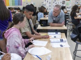 NASA STEM Initiatives Inspire Learning Outside School A group of students sitting at a desk in a classroom work on constructing paper airplanes. A man leans on the desk, speaking with the students. Behind the man are other students seated at desks.