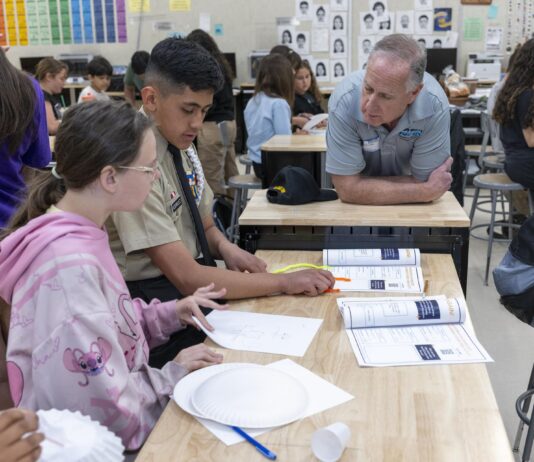 NASA STEM Initiatives Inspire Learning Outside School A group of students sitting at a desk in a classroom work on constructing paper airplanes. A man leans on the desk, speaking with the students. Behind the man are other students seated at desks.