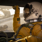 NASA ER-2 Pilot Readies for GEMx Mission Flight A person facing left sits inside a vehicle. They are wearing a yellow jumpsuit and gloves and a white helmet with a visor that looks like a smaller version of the kind astronauts wear during spacewalks. Outside of the window behind the person, you can see the ER-2 aircraft being prepared for flight.