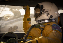 NASA ER-2 Pilot Readies for GEMx Mission Flight A person facing left sits inside a vehicle. They are wearing a yellow jumpsuit and gloves and a white helmet with a visor that looks like a smaller version of the kind astronauts wear during spacewalks. Outside of the window behind the person, you can see the ER-2 aircraft being prepared for flight.