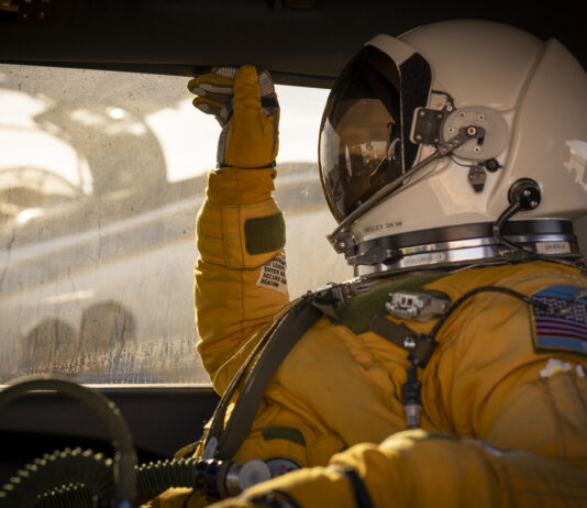 NASA ER-2 Pilot Readies for GEMx Mission Flight A person facing left sits inside a vehicle. They are wearing a yellow jumpsuit and gloves and a white helmet with a visor that looks like a smaller version of the kind astronauts wear during spacewalks. Outside of the window behind the person, you can see the ER-2 aircraft being prepared for flight.
