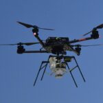 NASA Experiments with Supersonic Parachute Delivery System A drone with four rotors hoovers against a canvas of deep blue sky prior to releasing the experiment it carries high above the desert floor.