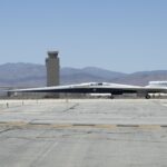 NASA’s X-59 Supersonic Jet Initiates Ground Taxi Trials NASA’s X-59 aircraft taxis on the runway at U.S. Air Force Plant 42 in Palmdale, California. The aircraft is shown in profile moving from right to left, with the control tower and desert mountains in the background. In the foreground, the gray concrete runway features a solid yellow line with evenly spaced parallel black stripes inside it, all under a clear blue sky.