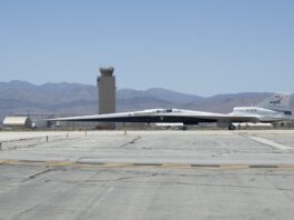 NASA’s X-59 Supersonic Jet Initiates Ground Taxi Trials NASA’s X-59 aircraft taxis on the runway at U.S. Air Force Plant 42 in Palmdale, California. The aircraft is shown in profile moving from right to left, with the control tower and desert mountains in the background. In the foreground, the gray concrete runway features a solid yellow line with evenly spaced parallel black stripes inside it, all under a clear blue sky.