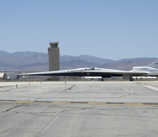 NASA’s X-59 Supersonic Jet Initiates Ground Taxi Trials NASA’s X-59 aircraft taxis on the runway at U.S. Air Force Plant 42 in Palmdale, California. The aircraft is shown in profile moving from right to left, with the control tower and desert mountains in the background. In the foreground, the gray concrete runway features a solid yellow line with evenly spaced parallel black stripes inside it, all under a clear blue sky.