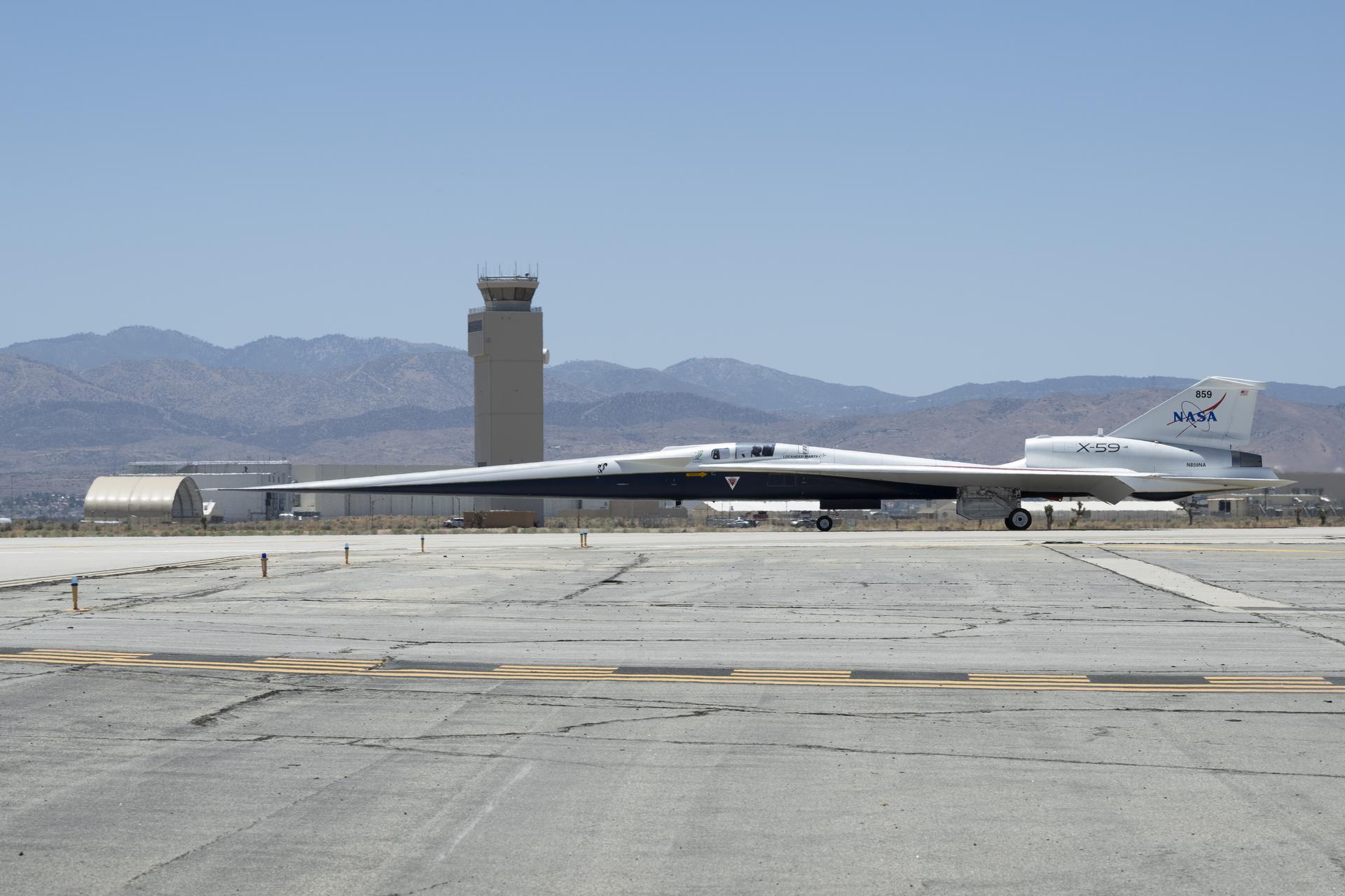 afrc2025 0112 06large.jpg NASA’s X-59 aircraft taxis on the runway at U.S. Air Force Plant 42 in Palmdale, California. The aircraft is shown in profile moving from right to left, with the control tower and desert mountains in the background. In the foreground, the gray concrete runway features a solid yellow line with evenly spaced parallel black stripes inside it, all under a clear blue sky.