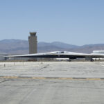 NASA’s X-59 Aircraft Relocates for Upcoming Developments NASA’s X-59 quiet supersonic research aircraft taxis across the runway at U.S. Air Force Plant 42. The aircraft has a distinctive, long and pointy nose. An air control tower is visible behind the plane. Even further in the distance beneath a hazy blue sky are hills and mountains.