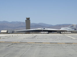 NASA’s X-59 Aircraft Relocates for Upcoming Developments NASA’s X-59 quiet supersonic research aircraft taxis across the runway at U.S. Air Force Plant 42. The aircraft has a distinctive, long and pointy nose. An air control tower is visible behind the plane. Even further in the distance beneath a hazy blue sky are hills and mountains.