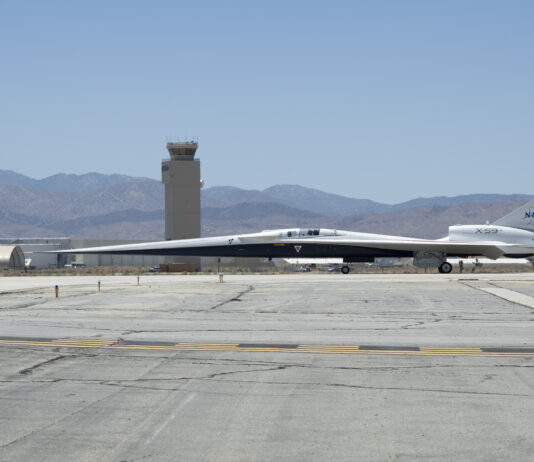 NASA’s X-59 Aircraft Relocates for Upcoming Developments NASA’s X-59 quiet supersonic research aircraft taxis across the runway at U.S. Air Force Plant 42. The aircraft has a distinctive, long and pointy nose. An air control tower is visible behind the plane. Even further in the distance beneath a hazy blue sky are hills and mountains.