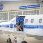 NASA PC-12 aircraft aids flight research agency-wide. The left side of a white aircraft with one door and three windows sits on a concrete ramp. The aircraft is painted with a blue stripe and a round NASA logo on the side. A man in a blue jacket and tan pants climbs out of the aircraft carrying two black bags. A silver hangar sits in the background with large white doors. A round NASA logo is located on the hangar as well as a blue sign with white letters to the right side.