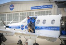 NASA PC-12 aircraft aids flight research agency-wide. The left side of a white aircraft with one door and three windows sits on a concrete ramp. The aircraft is painted with a blue stripe and a round NASA logo on the side. A man in a blue jacket and tan pants climbs out of the aircraft carrying two black bags. A silver hangar sits in the background with large white doors. A round NASA logo is located on the hangar as well as a blue sign with white letters to the right side.