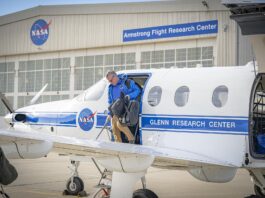 NASA PC-12 aircraft aids flight research agency-wide. The left side of a white aircraft with one door and three windows sits on a concrete ramp. The aircraft is painted with a blue stripe and a round NASA logo on the side. A man in a blue jacket and tan pants climbs out of the aircraft carrying two black bags. A silver hangar sits in the background with large white doors. A round NASA logo is located on the hangar as well as a blue sign with white letters to the right side.