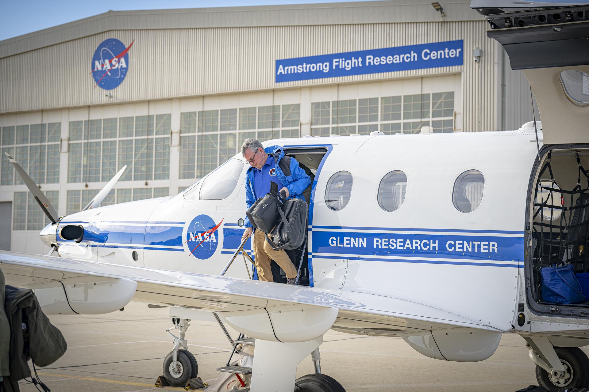 The left side of a white aircraft with one door and three windows sits on a concrete ramp. The aircraft is painted with a blue stripe and a round NASA logo on the side. A man in a blue jacket and tan pants climbs out of the aircraft carrying two black bags. A silver hangar sits in the background with large white doors. A round NASA logo is located on the hangar as well as a blue sign with white letters to the right side.