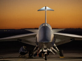 Second Flight Scheduled for NASA’s X-59 Aircraft An aircraft resting on a section of runway as seen from the front, with its nose facing the camera. The early morning sky is orange in the background.