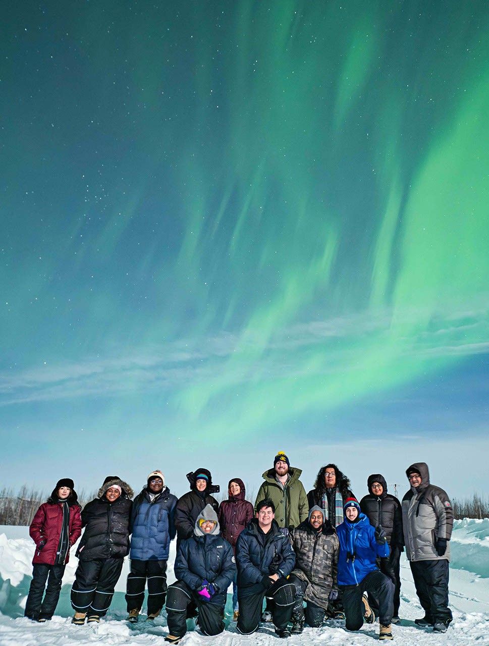A group of 13 people standing together on snow-covered ground, dressed in warm winter clothing, with the aurora glowing in the sky above them. Some are kneeling, while others stand in front of them, all facing the camera and smiling.