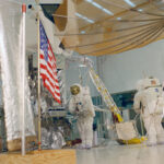 Remembering 1968: Apollo 13, 14 Prep and Apollo 12 Tour Two astronauts wearing white spacesuits practice a moonwalk, with an American flag visible in front of a lunar module mockup.