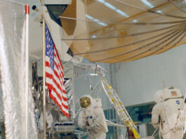 Remembering 1968: Apollo 13, 14 Prep and Apollo 12 Tour Two astronauts wearing white spacesuits practice a moonwalk, with an American flag visible in front of a lunar module mockup.