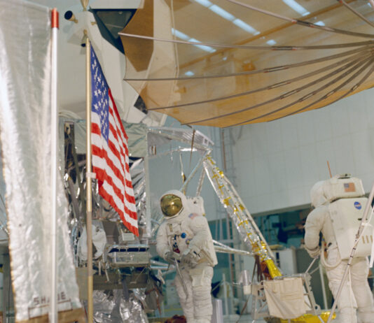 Remembering 1968: Apollo 13, 14 Prep and Apollo 12 Tour Two astronauts wearing white spacesuits practice a moonwalk, with an American flag visible in front of a lunar module mockup.