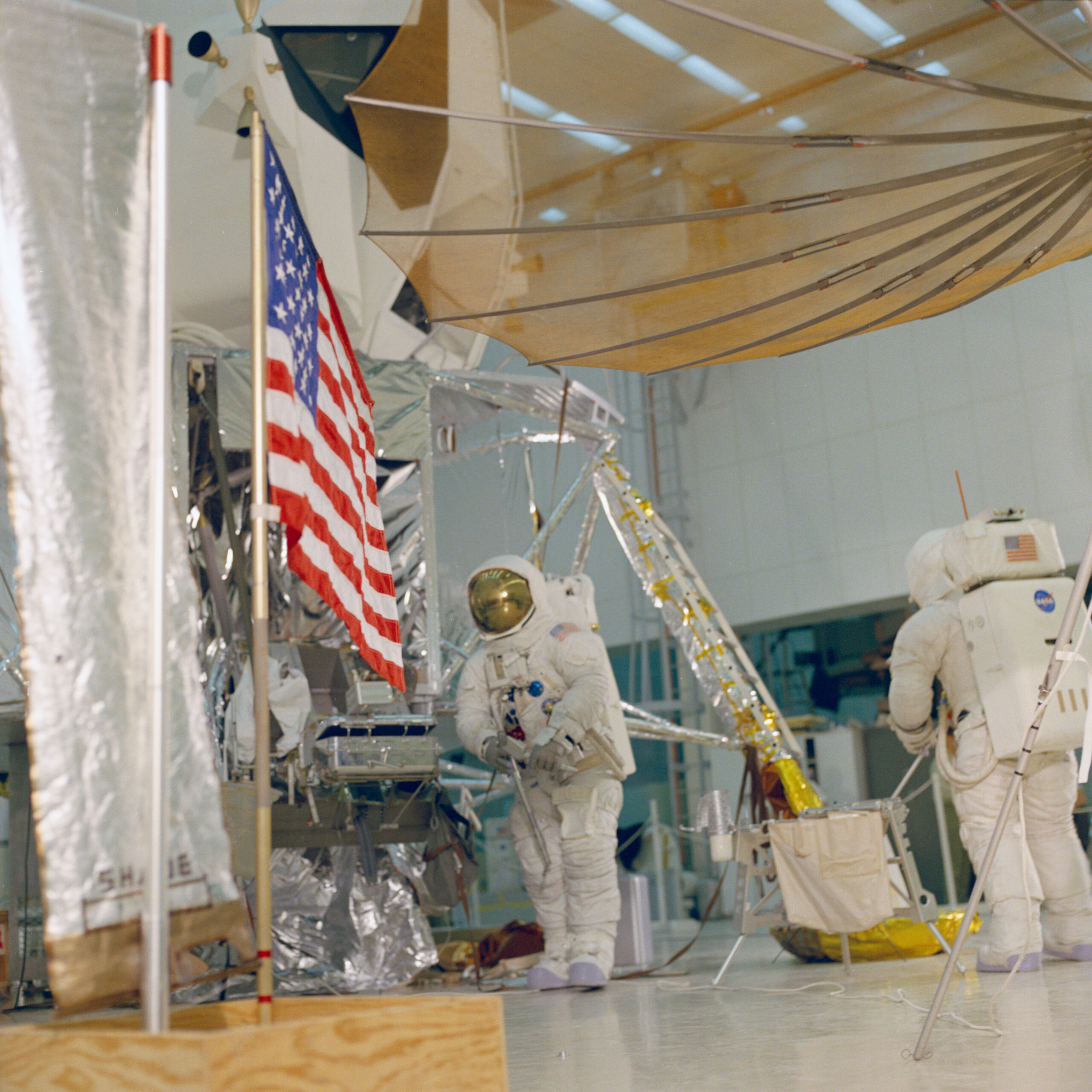 apollo 13 l 2 months 6 eva training lovell haise ksc feb 12 1970 s70 27199.jpg Two astronauts wearing white spacesuits practice a moonwalk, with an American flag visible in front of a lunar module mockup.