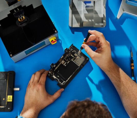 Apple expands repair options by supporting genuine used parts Technician repairing smartphone on blue workbench.
