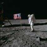NASA Celebrates 55 Years Since Apollo 11 Moon Landing On the desolate pock-marked lunar surface, astronaut Edwin E. Aldrin Jr., wearing a white Extravehicular Mobility Unit spacesuit, stands to the right of the American flag planted in the soil. The flag is unfurled and waving to the left, with Aldrin facing it in the image and seen from a side view.
