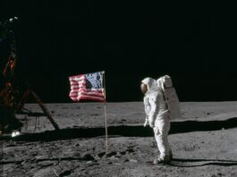 NASA Celebrates 55 Years Since Apollo 11 Moon Landing On the desolate pock-marked lunar surface, astronaut Edwin E. Aldrin Jr., wearing a white Extravehicular Mobility Unit spacesuit, stands to the right of the American flag planted in the soil. The flag is unfurled and waving to the left, with Aldrin facing it in the image and seen from a side view.