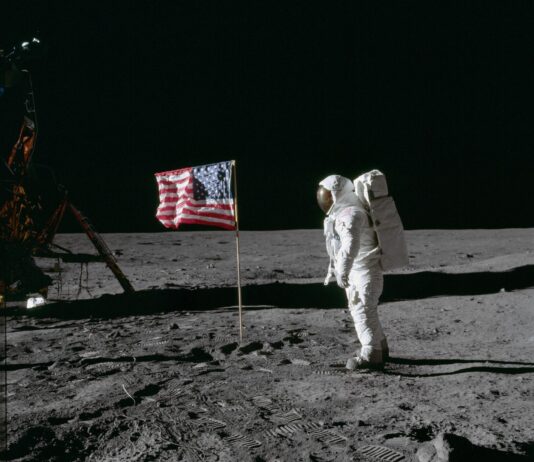 NASA Celebrates 55 Years Since Apollo 11 Moon Landing On the desolate pock-marked lunar surface, astronaut Edwin E. Aldrin Jr., wearing a white Extravehicular Mobility Unit spacesuit, stands to the right of the American flag planted in the soil. The flag is unfurled and waving to the left, with Aldrin facing it in the image and seen from a side view.