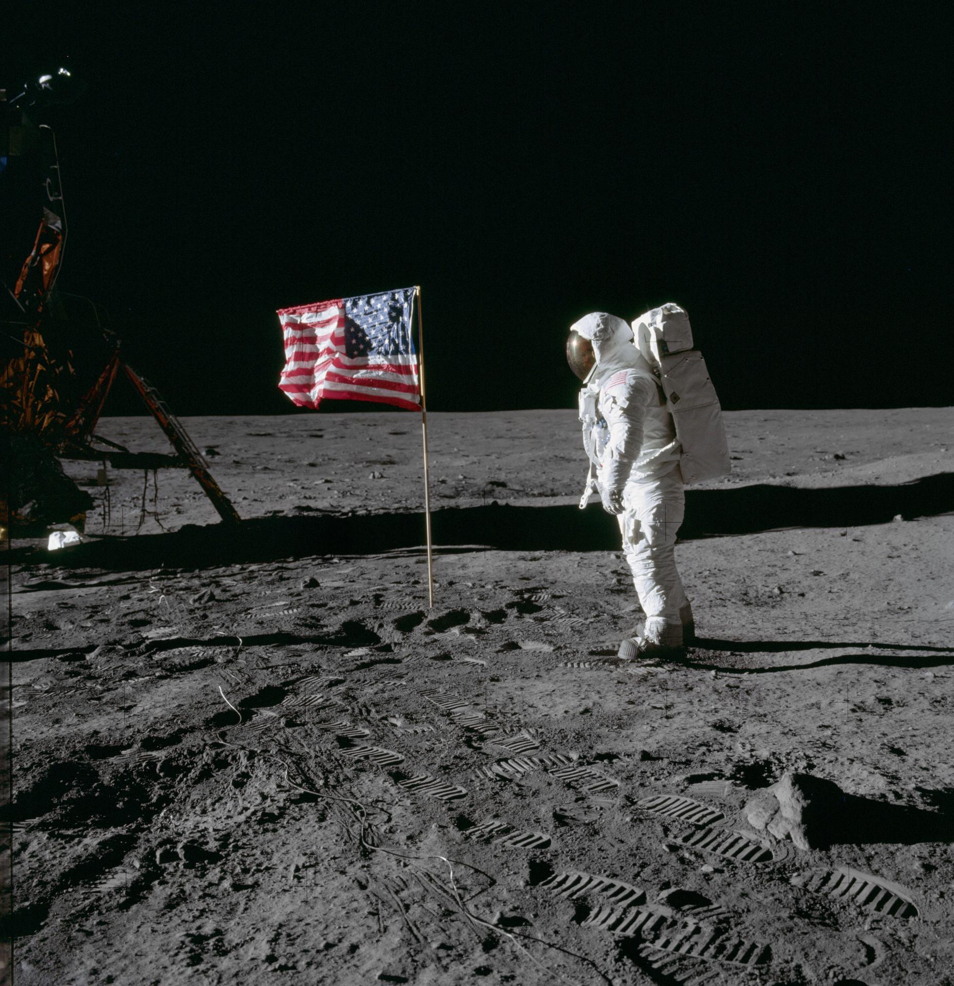 as11 40 5875large.jpg On the desolate pock-marked lunar surface, astronaut Edwin E. Aldrin Jr., wearing a white Extravehicular Mobility Unit spacesuit, stands to the right of the American flag planted in the soil. The flag is unfurled and waving to the left, with Aldrin facing it in the image and seen from a side view.